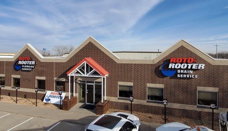 The exterior of a brick Roto-Rooter building under a bright blue sky with parts of two cars visible in the foreground.