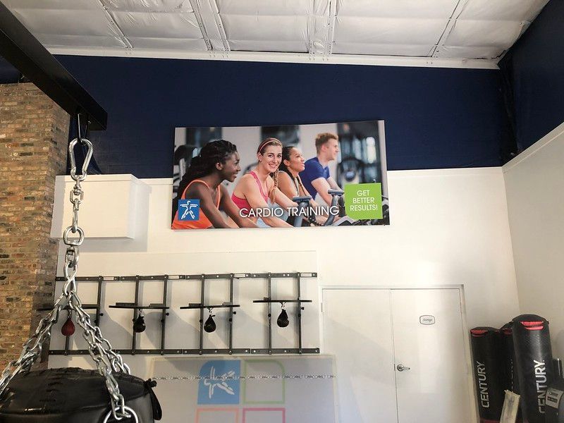 A gym wall features a promotional banner of people exercising, with a heavy bag and equipment in the foreground.