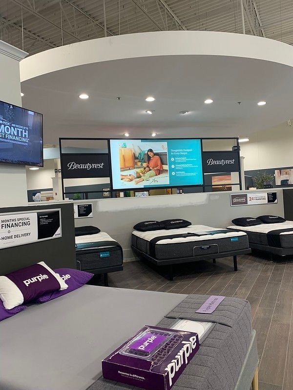 A Purple mattress store showroom with multiple beds on display under bright lights and a large promotional screen.