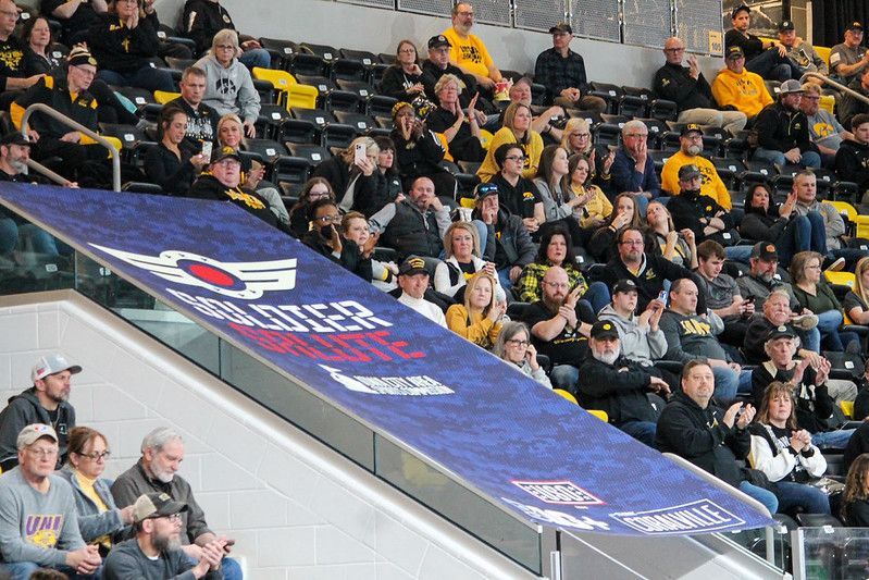 A crowd of spectators watches a sporting event from arena seating, beside a large blue banner with team branding.