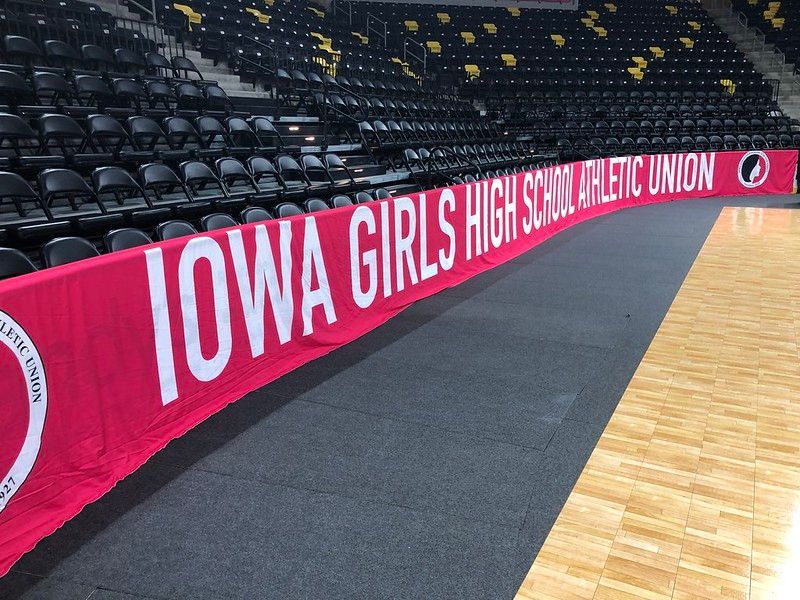 A red banner for the Iowa Girls High School Athletic Union lining the sideline of an indoor basketball court.