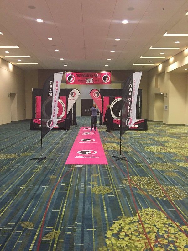 A pink carpeted path leads toward two people standing before an event banner and branded displays in a hotel ballroom.