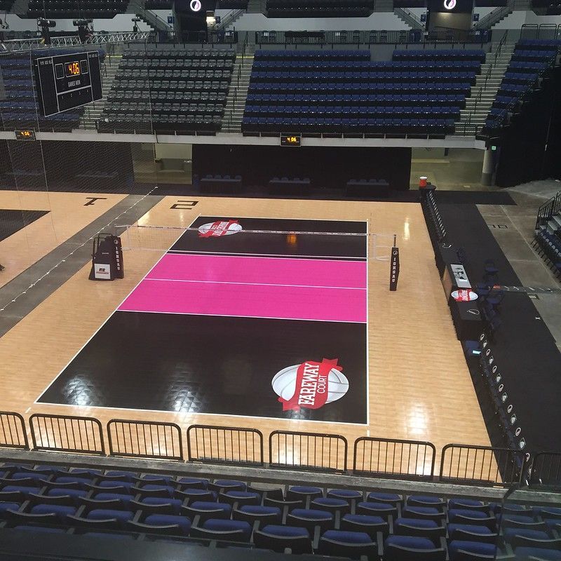An empty indoor volleyball court featuring a bright pink center, surrounded by stadium seating in a darkened arena.