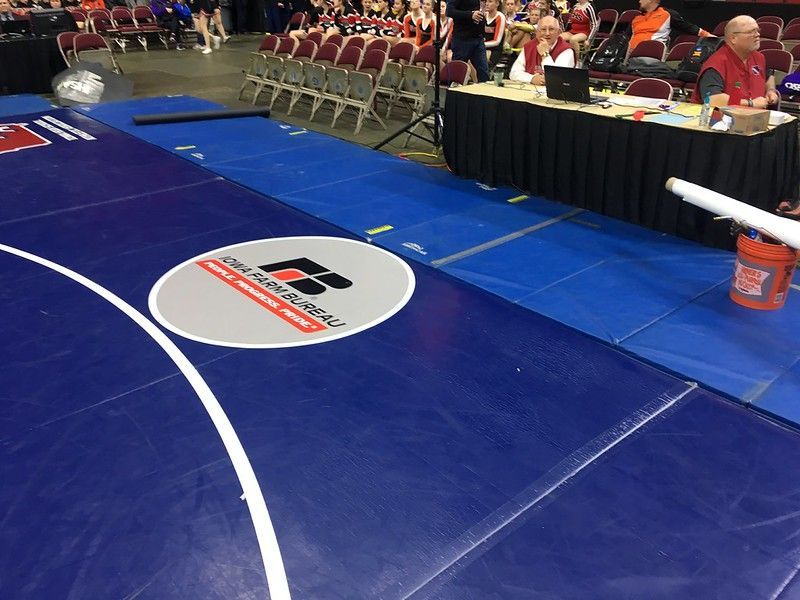 A blue wrestling mat with a logo in the center and a table with officials in the background.