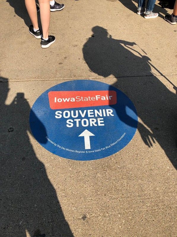 A circular blue Iowa State Fair Souvenir Store sign with a white arrow on the ground, surrounded by people's feet.