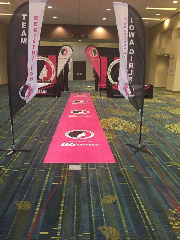 A pink runner carpet leads to a conference room entrance flanked by two tall black feather banners with white text.