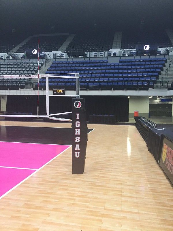 A volleyball court with a net and an IGHSAU-branded pole, featuring a pink floor and empty blue stadium seating.