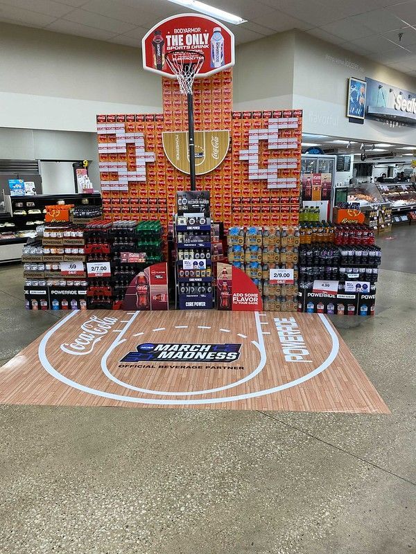 A March Madness-themed Coca-Cola display in a grocery store featuring a cardboard basketball hoop and court floor decal.