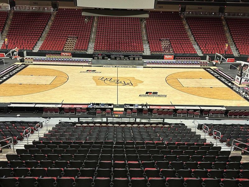 An empty basketball arena featuring a light wood court, black seating, and a large red-seated stadium backdrop.