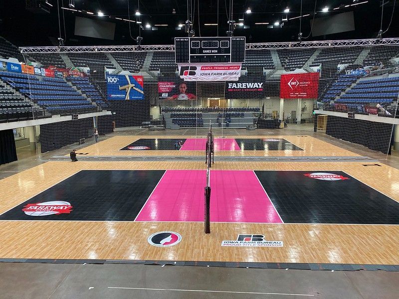 An indoor volleyball arena with two wooden courts featuring black and vibrant pink sections and various sponsor logos.