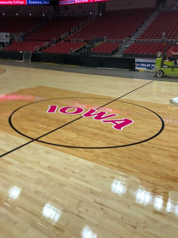 The Iowa Hawkeyes logo painted in pink on a basketball court, with red arena seating in the background.