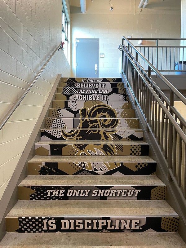 Stairs in a stairwell with gold and black patterns, a ram mascot, and motivational text about belief and discipline.