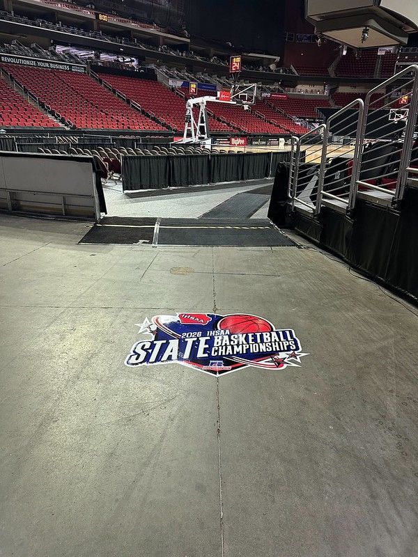 A basketball arena floor with a large “State Basketball Championships” decal, featuring red seats in the background.