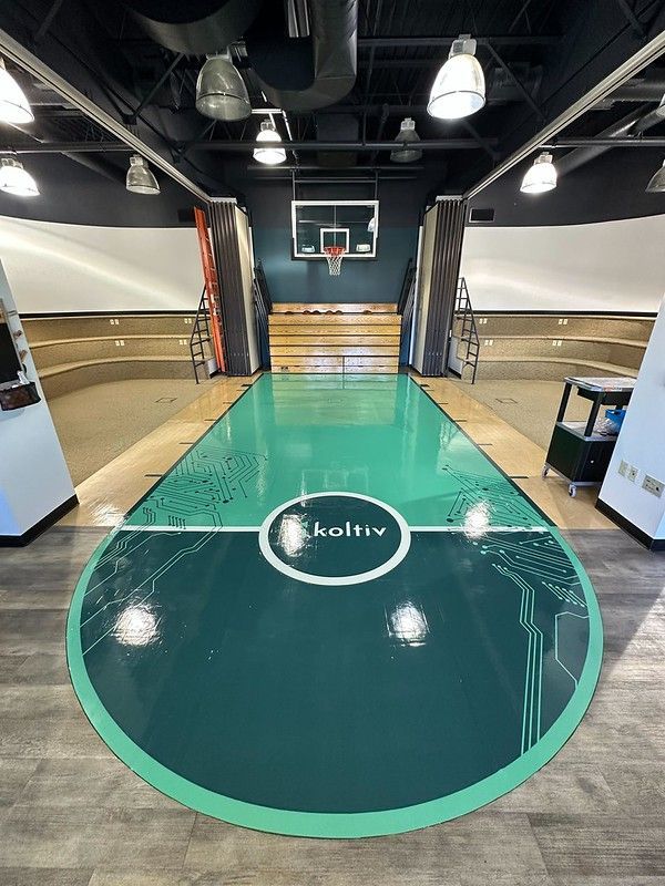 Indoor basketball court with a green floor, Koltiv logo, and tiered wooden stadium seating under industrial lighting.
