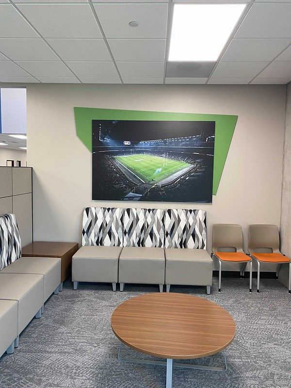 A waiting area with beige and patterned modular seating, a round wood table, and stadium art on a green wall accent.