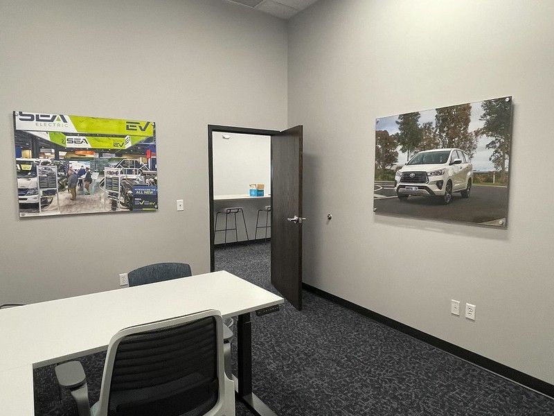 An office with a desk, two chairs, and two framed car photos on the light gray walls, with an open door to a break room.