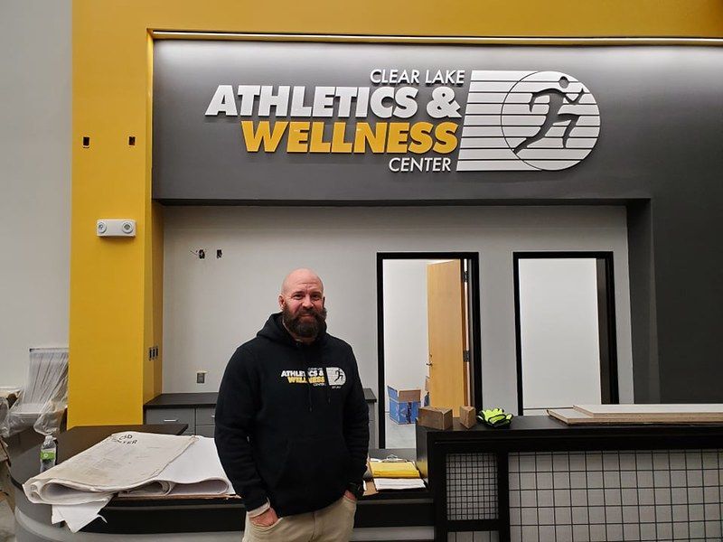 A person stands at a front desk inside the Clear Lake Athletics & Wellness Center, which has yellow and grey walls.