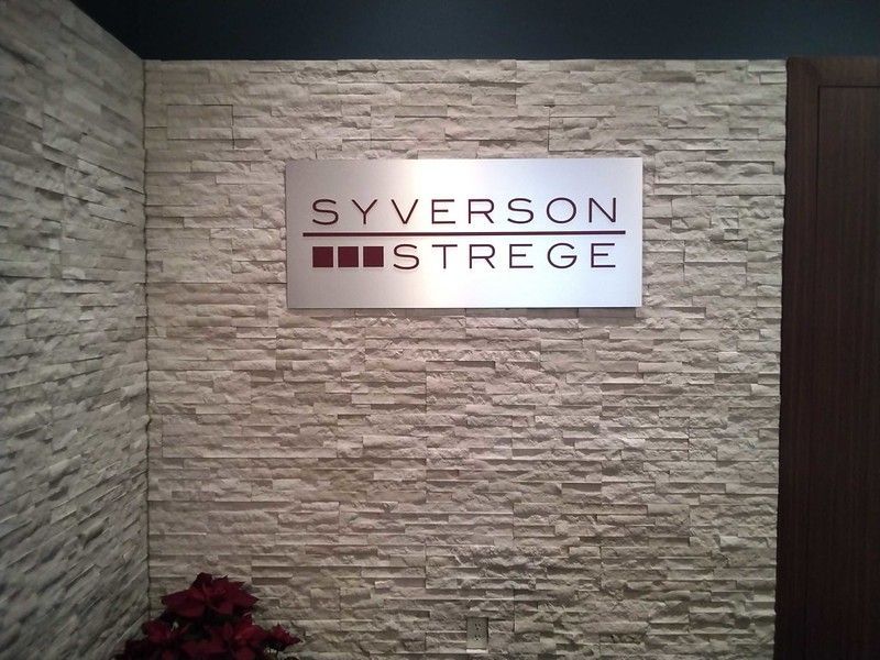 The Syverson Strege company logo on a rectangular metal sign mounted to a light-colored, textured stone wall.