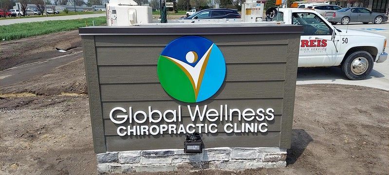 A monument sign for Global Wellness Chiropractic Clinic with a blue, green, and white circular logo on a dark brown wall.