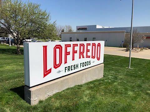 A white rectangular sign for Loffredo Fresh Foods with red lettering on a grassy lawn in front of a warehouse building.