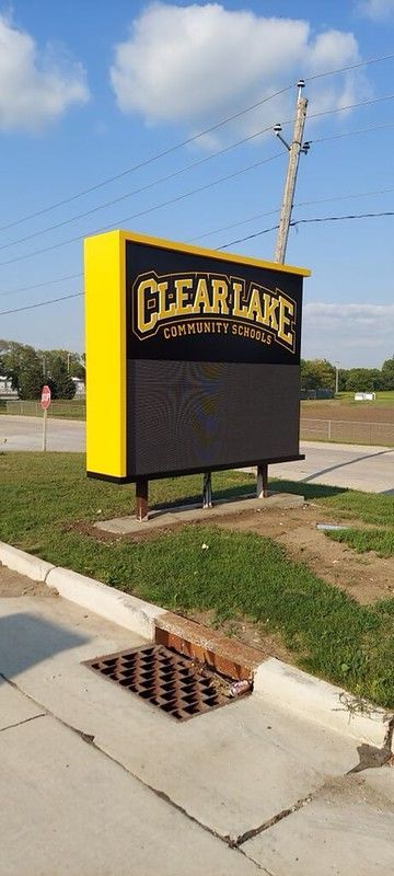 A yellow and black Clear Lake sign stands on a grassy area next to a sidewalk with a storm drain.