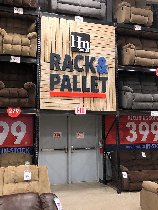 A furniture warehouse display featuring rows of reclining sofas on metal racks surrounding a central set of exit doors.