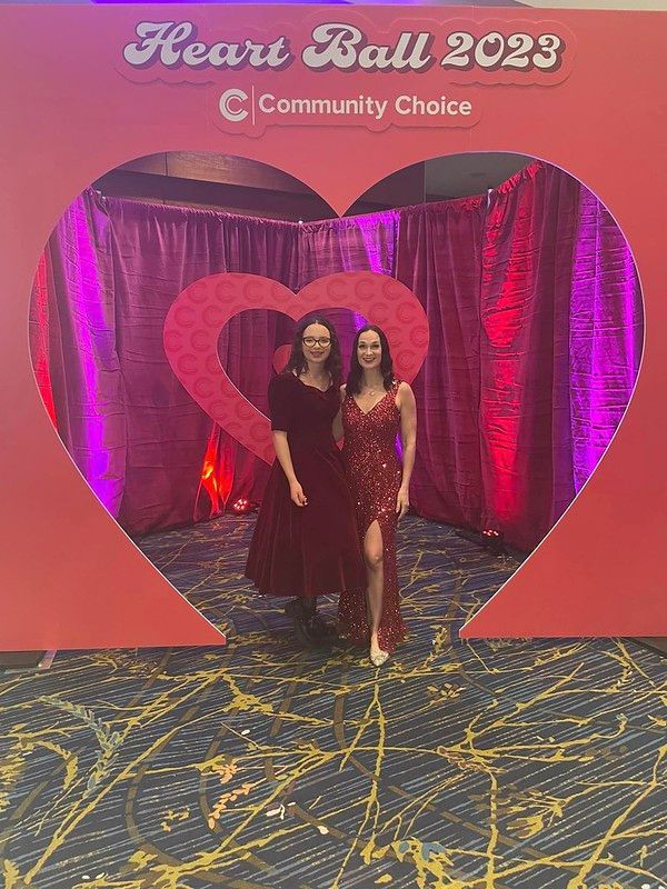 Two people pose inside a large red heart cutout at the 2023 Heart Ball, framed by maroon curtains on a patterned carpet.