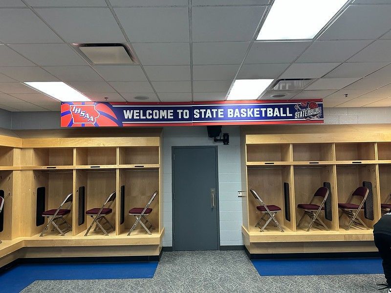 A locker room features wooden cubbies, folding chairs, and a red, white, and blue banner reading 