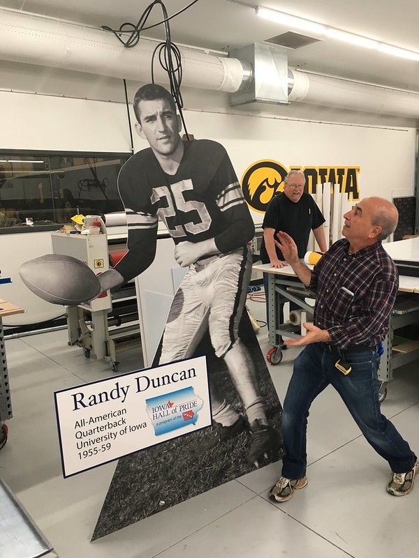 A person stands beside a life-size cutout of football player Randy Duncan in a workshop with Iowa branding.