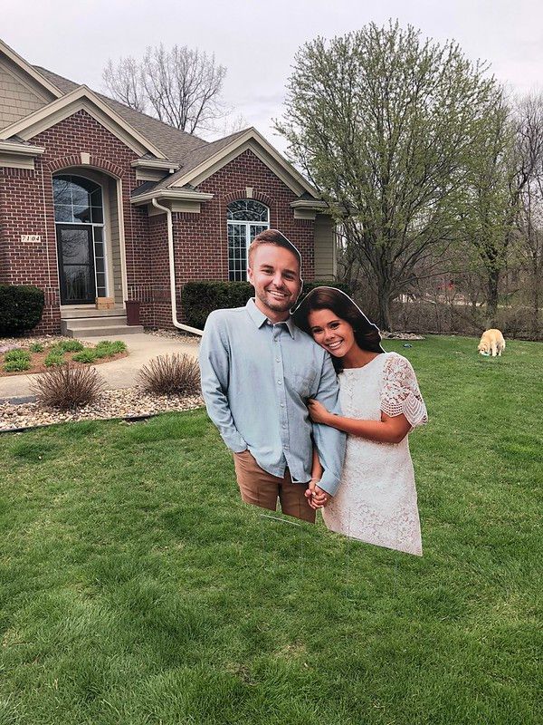 A man in a light blue shirt and a woman in a white dress stand smiling together on a grassy lawn in front of a brick home.