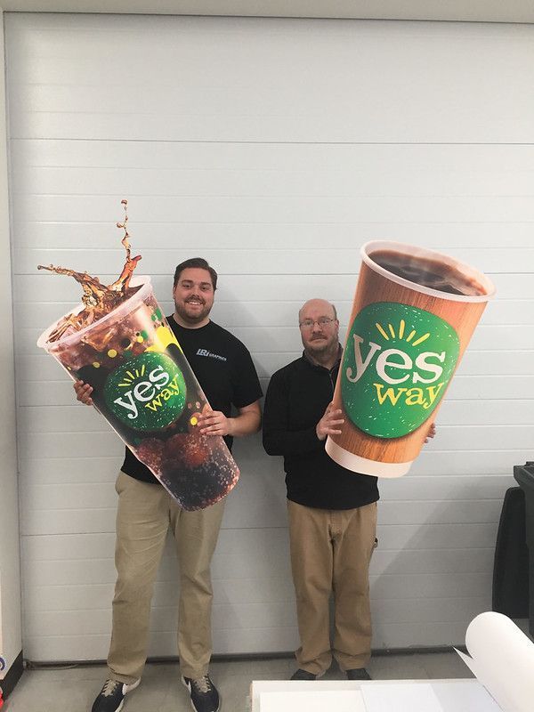 Two people stand side-by-side in front of a white wall, each holding a large cardboard cutout of a Yesway branded cup.