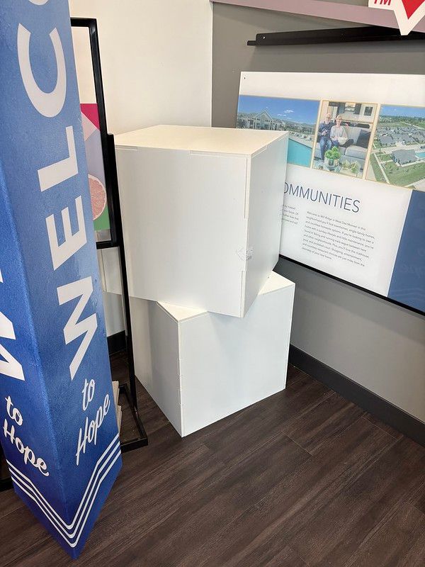 Two stacked white square pedestals stand on a dark floor in a room next to a welcome sign and a wall display of communities.
