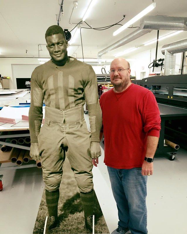 A person in a red shirt stands next to a life-size cutout of a man in vintage athletic gear in a workshop.