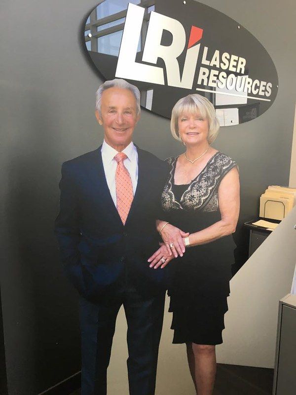 A man in a suit and a woman in a black dress stand smiling in front of a Laser Resources office wall sign.