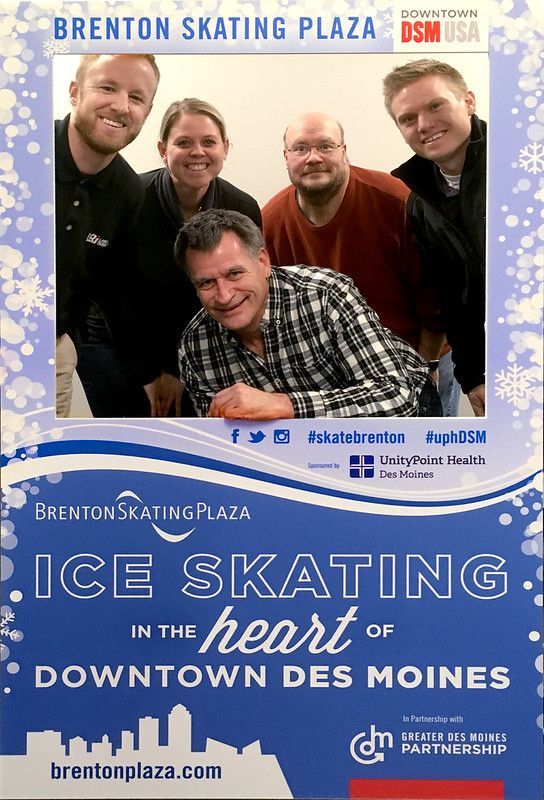 Five people pose behind a Brenton Skating Plaza sign for ice skating in downtown Des Moines.