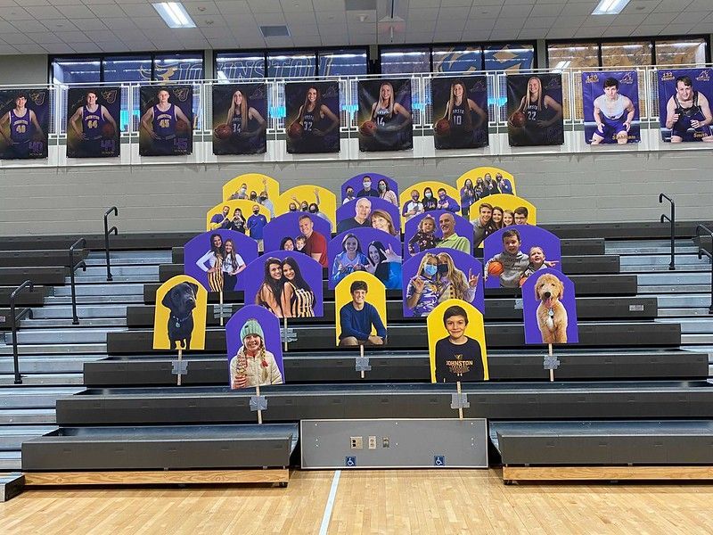 Cutout portraits and photos of people and pets are arranged on gymnasium bleachers, below athletic banners on the wall.