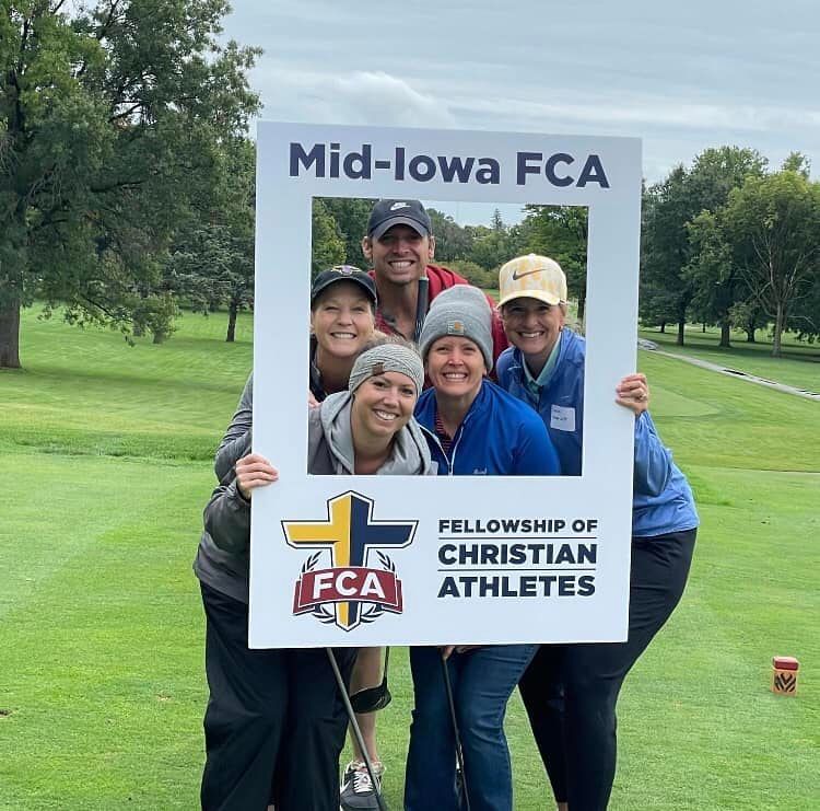 Five people smiling while holding a Mid-Iowa FCA photo frame on a golf course.