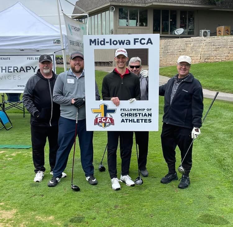 Four people stand on a grassy golf course holding a Mid-Iowa Fellowship of Christian Athletes sign for a group photo.