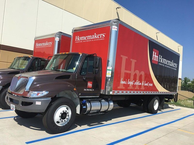 Two brown Homemakers furniture delivery trucks parked in a lot under a clear blue sky.