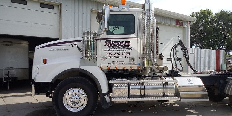 A white semi-truck parked in front of a warehouse building, featuring chrome accents and company branding on the door.