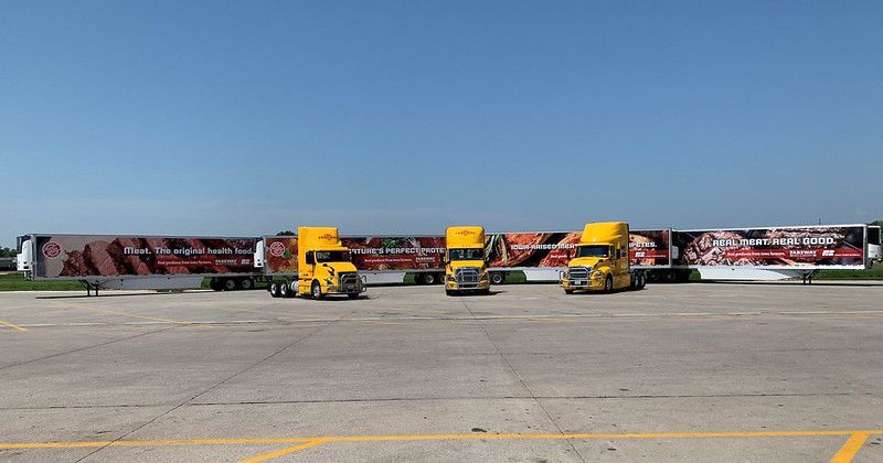 Three yellow semi-trucks with matching branded trailers parked side-by-side on an expansive concrete lot under a clear sky.