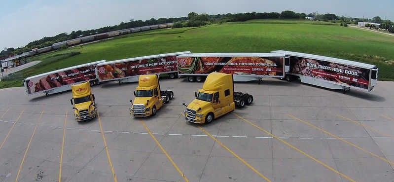 Three yellow semi-trucks with matching branded trailers parked in a row on a paved lot with a grassy field in the background.