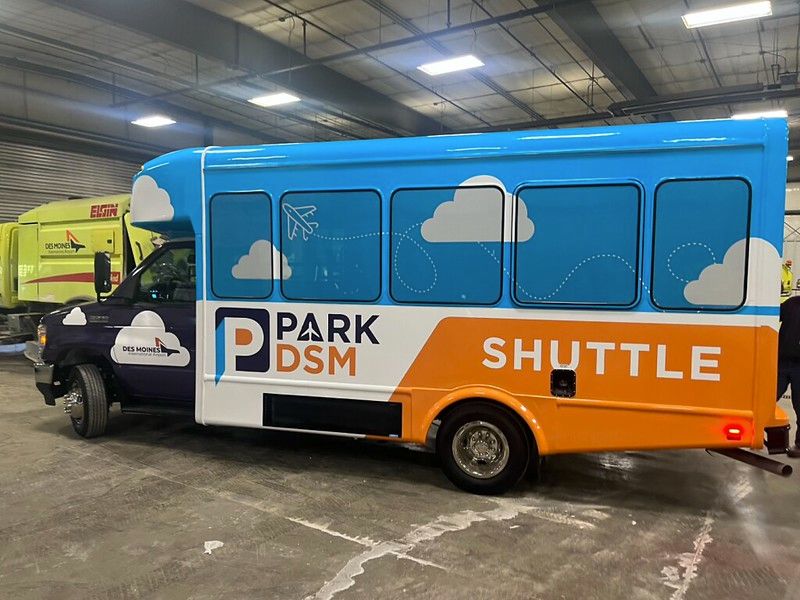 A Park DSM blue and orange shuttle bus parked inside a warehouse with a plane and cloud graphic on the side.