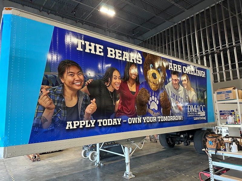 A promotional advertisement on a semi-trailer for DMACC featuring smiling people and a bear mascot with text.