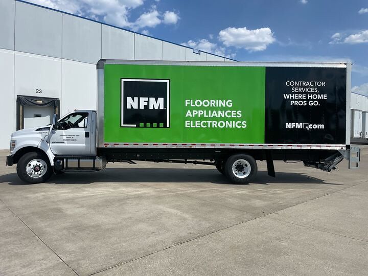 A silver NFM box truck parked in a concrete lot, with green and black branding advertising flooring and appliances.