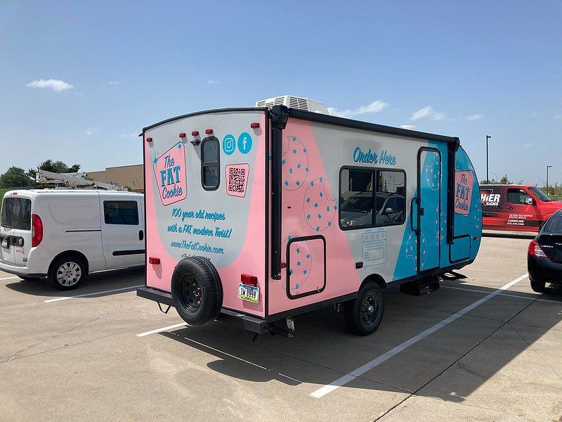 A Fat Cookie food trailer with pink and blue geometric patterns parked in an outdoor lot on a sunny day.