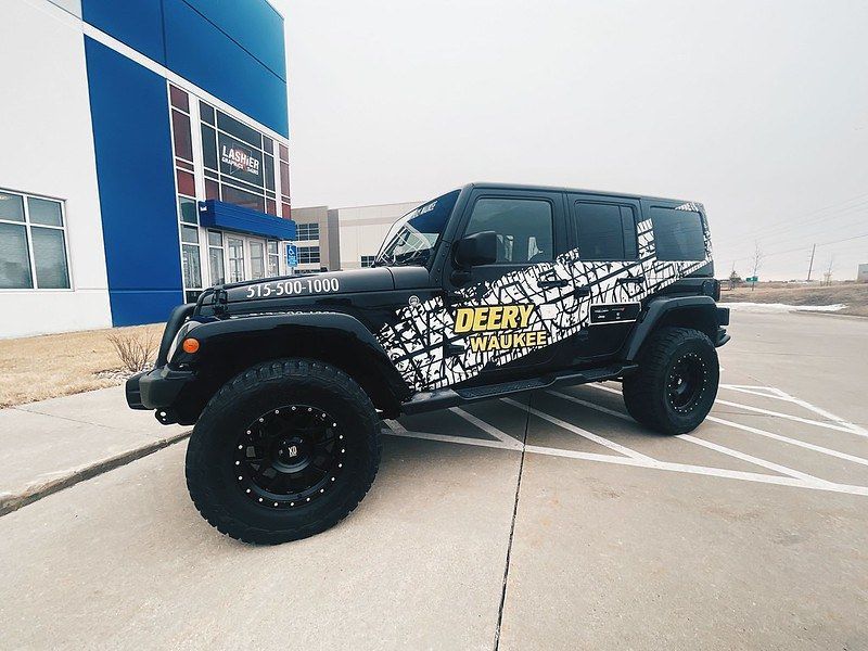 A black Jeep Wrangler with custom white tire-tread graphics parked in front of a building with blue and white accents.