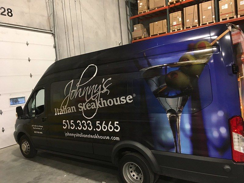 A side view of a black van wrapped with the Johnny's Italian Steakhouse logo, contact information, and a martini graphic.