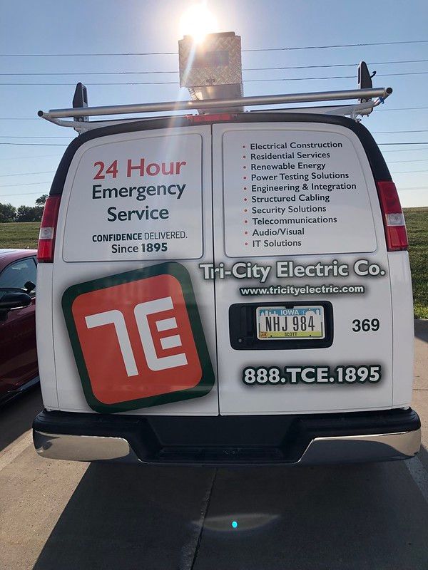 The back of a white Tri-City Electric Co. work van with their logo, contact information, and service list on a sunny road.