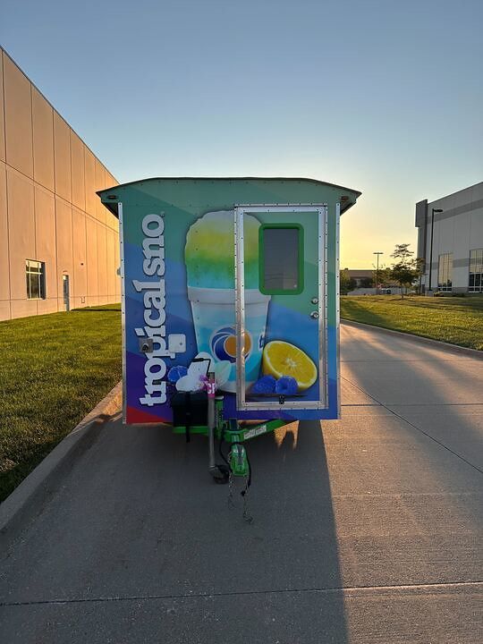 A colorful Tropical Sno trailer parked on a paved road next to buildings at sunset.